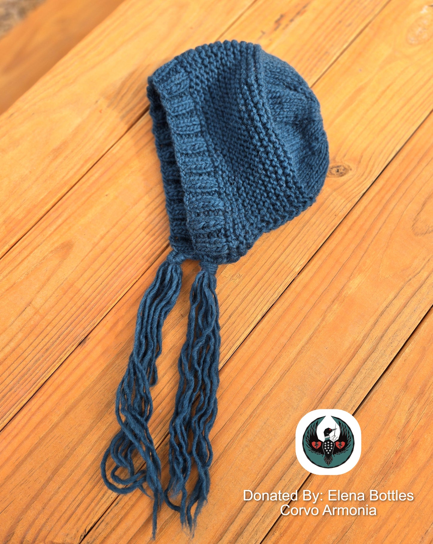 teal knitted hat with tassels on a wooden surface, donated by Elena Bottles Corvo Armonia.