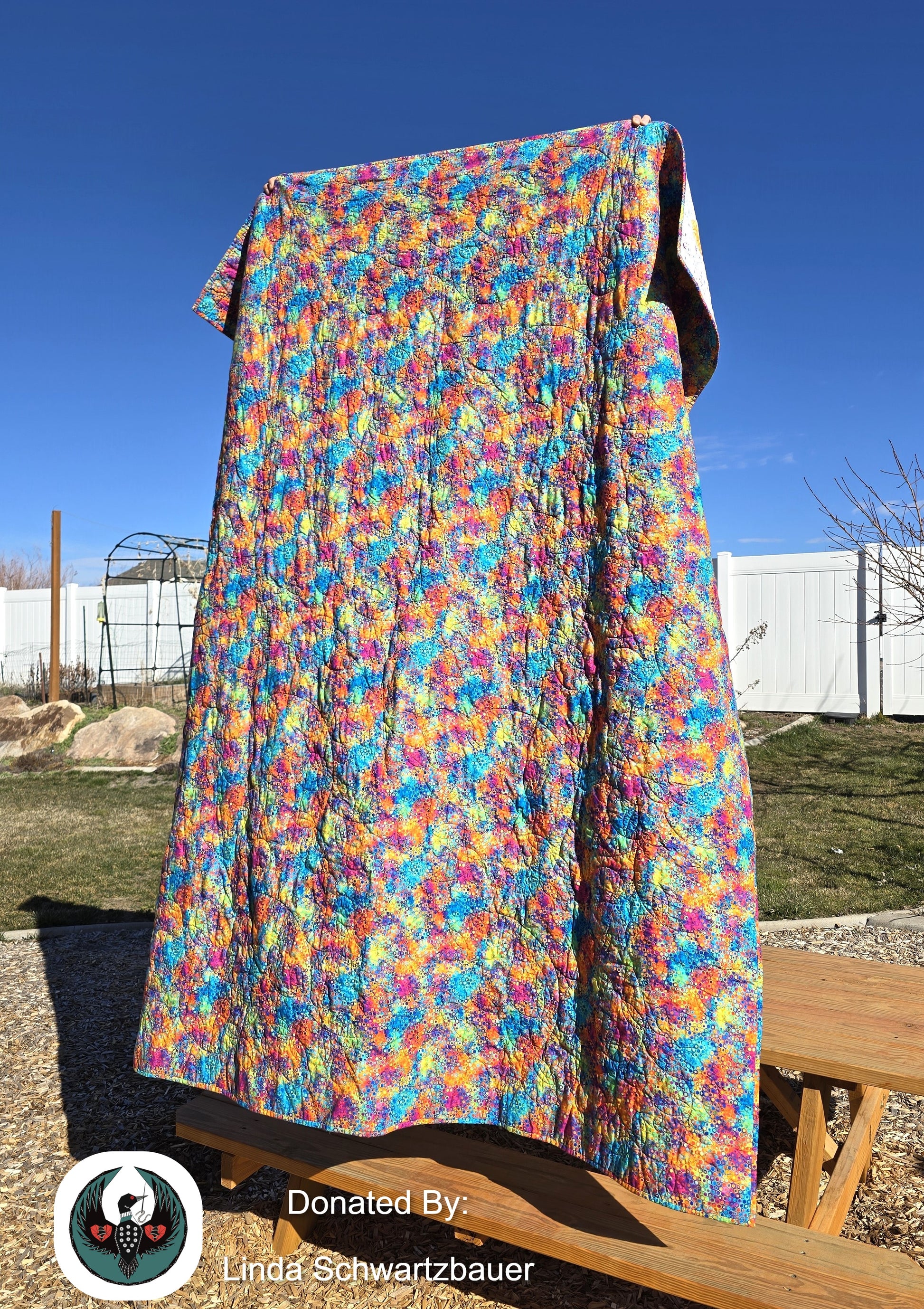 Rainbow quilt in vibrant colors held up outdoors against a blue sky.