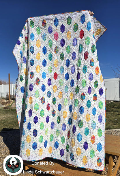 Rainbow quilt with a geometric hexagon pattern held up outdoors against a blue sky.