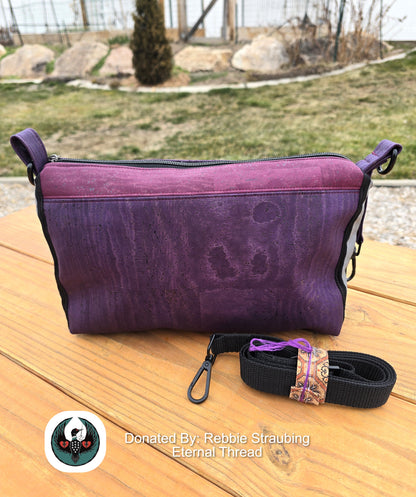 purple and eggplant handbag with a black crossbody strap sitting on a wooden table outdoors