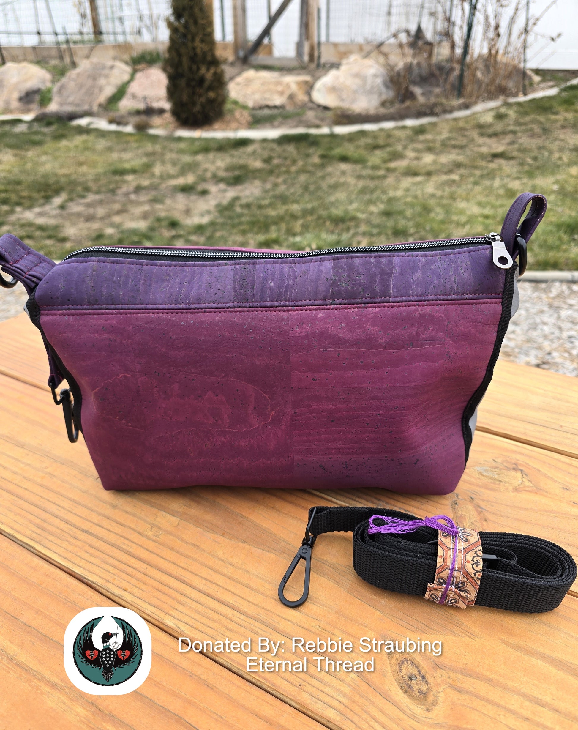 purple and eggplant handbag with a black crossbody strap sitting on a wooden table outdoors