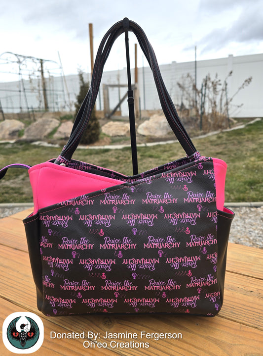 Black and pink tote bag with 'Raise the Matriarchy' text on a wooden surface outdoors.