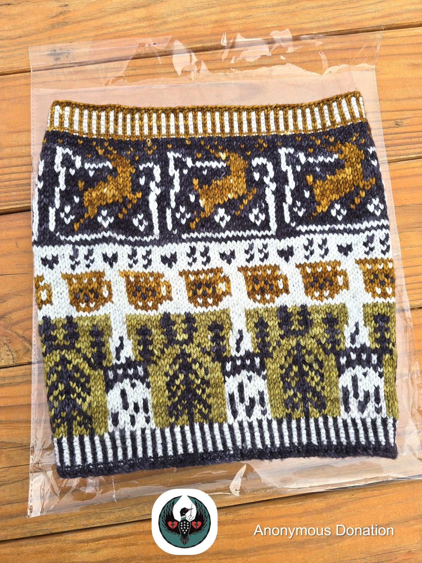 Knitted cowl in clear packaging on a wooden surface with 'Anonymous Donation' logo. Cowl has a design that features leaping deer, coffee mugs, and bridge towers