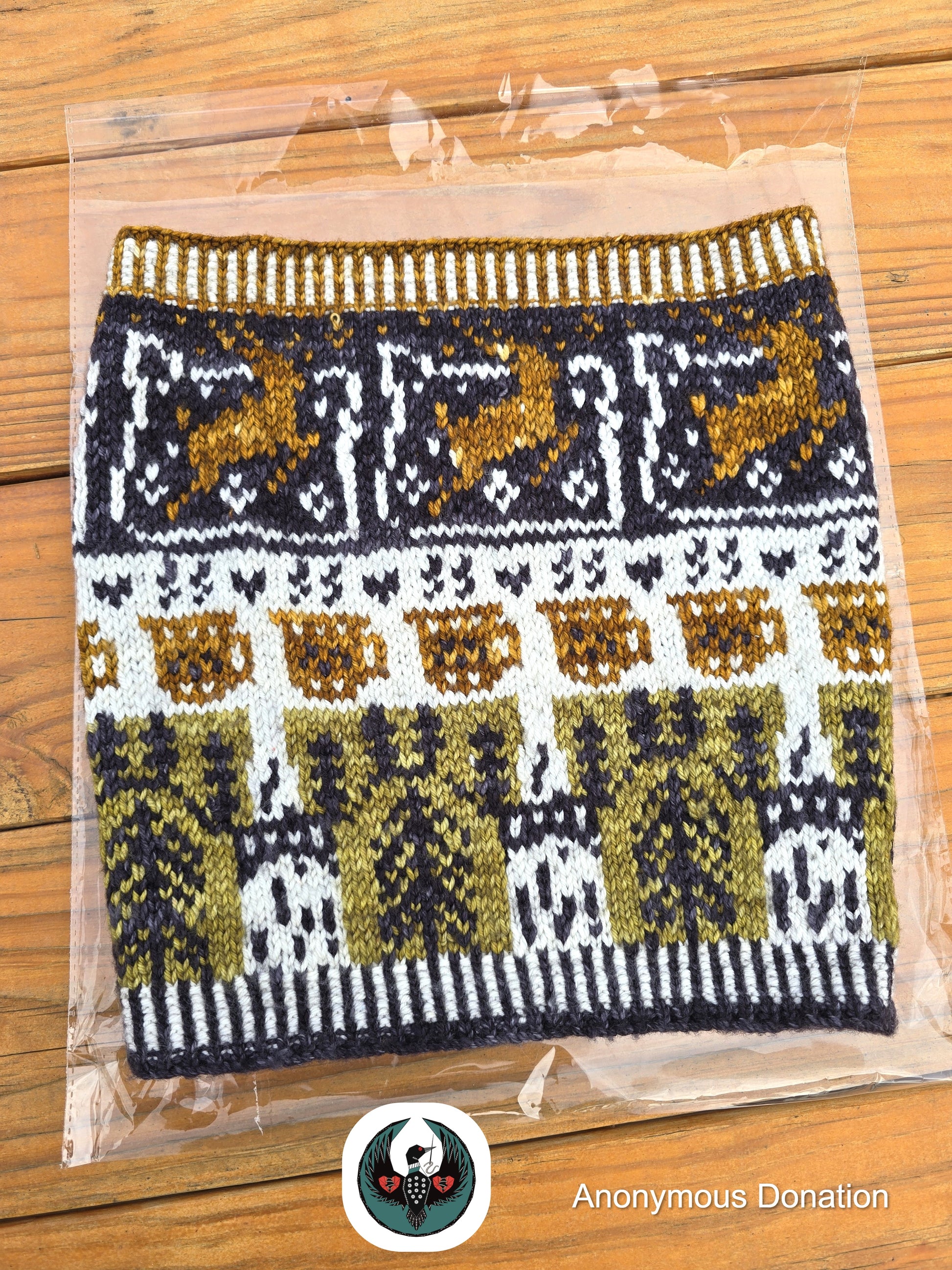 Knitted cowl in clear packaging on a wooden surface with 'Anonymous Donation' logo. Cowl has a design that features leaping deer, coffee mugs, and bridge towers