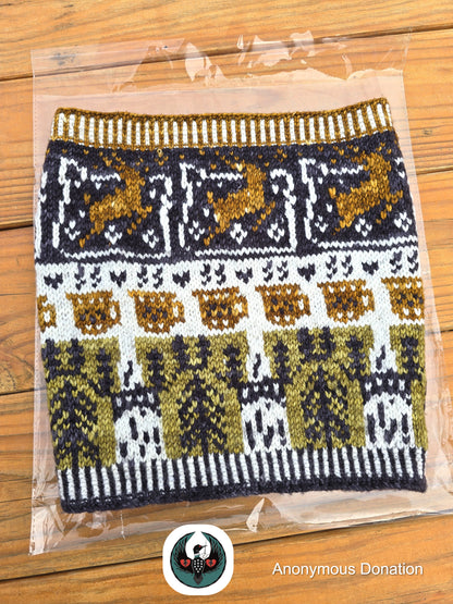 Knitted cowl in clear packaging on a wooden surface with 'Anonymous Donation' logo. Cowl has a design that features leaping deer, coffee mugs, and bridge towers