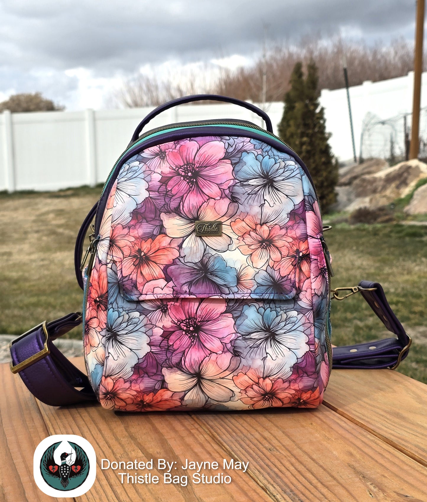 Floral patterned backpack on a wooden surface with a grassy area and trees in the background.