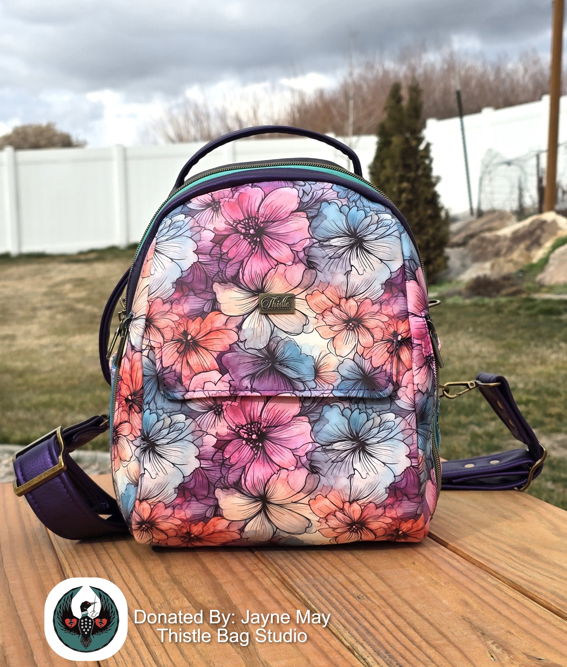 Floral patterned backpack on a wooden surface with a grassy area and trees in the background.