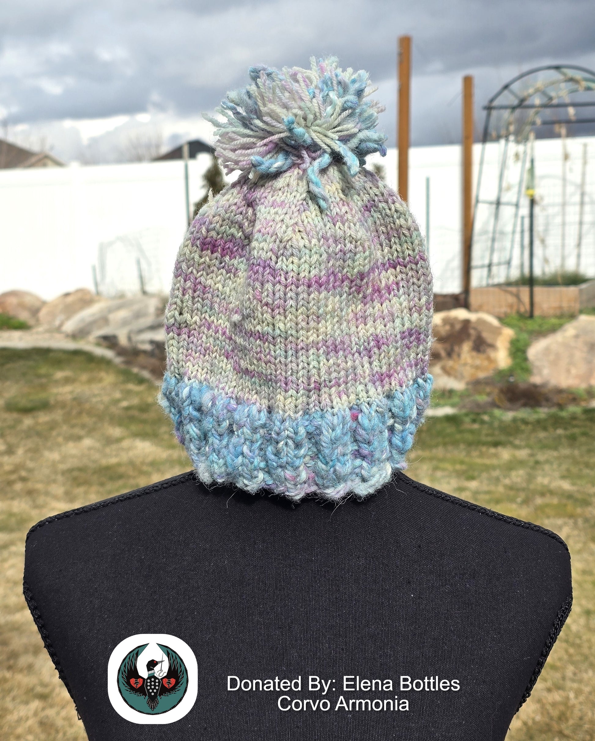 handknitted blue and pink beanie with a pompom top on a mannequin against an outdoor background.