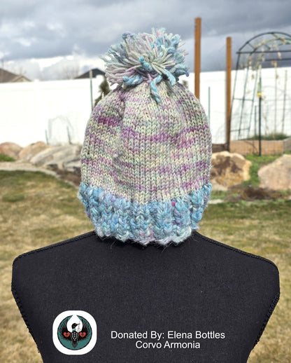 handknitted blue and pink beanie with a pompom top on a mannequin against an outdoor background.